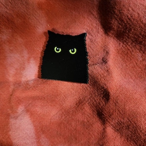 Puede incluir: Un pin de esmalte negro con forma de gato y ojos verde brillante. El pin tiene un contorno texturizado y está colocado sobre un fondo de tela roja. Los ojos del gato contrastan con el cuerpo negro.