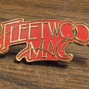 Puede incluir: Un broche de esmalte rojo con el texto Fleetwood Mac en color dorado y una tipografía estilizada. El broche tiene un borde de metal dorado que perfila la forma de las letras, creando un contraste definido con el fondo rojo vibrante.