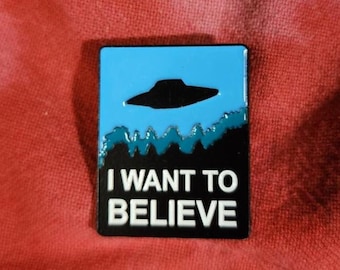 Puede incluir: Pin rectangular con un cielo azul y una silueta negra de OVNI. Debajo, un diseño de bosque negro y azul. El texto "I WANT TO BELIEVE" está en letras blancas sobre un fondo negro.