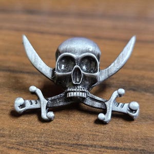 Puede incluir: Un emblema de calavera plateada con espadas cruzadas. La calavera presenta cuencas de los ojos detalladas, una cavidad nasal y dientes. Las espadas están colocadas detrás de la calavera, con las hojas curvadas hacia arriba. Probablemente sea un emblema o un objeto decorativo de temática pirata.