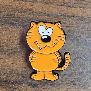May include: An orange enamel pin featuring a cartoon cat with a large head, wide white eyes, a black nose, and a single visible tooth. The cat has black outlines, striped markings on its body and tail, and a smiling expression with small whiskers.