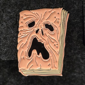 Könnte beinhalten: Ein Anstecknadel mit einem stilisierten, geöffneten Buch mit einem schreienden Gesicht auf dem Einband. Das Gesicht ist pfirsichfarben mit schwarzen Augen und Mund, und die Seiten sind hellgrün. Die Nadel ist wahrscheinlich aus Metall.