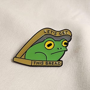 Puede incluir: Un pin de esmalte metálico que muestra una rana verde con grandes ojos amarillos asomándose desde dentro de una panera de color tostado. La tapa superior tiene el texto LET'S GET y la base inferior tiene el texto THIS BREAD en letras negras.