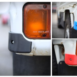 May include: Close-up of a white truck with a black plastic bumper guard. The bumper guard has a screw in the center. The truck has an orange turn signal lens. The truck is parked on a paved surface.