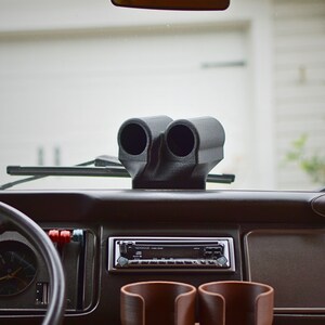 May include: A black dashboard with a double gauge pod, a car radio, and two cup holders. The gauge pod is mounted on the dashboard and has two circular openings for gauges. The radio is a standard car radio with a silver face and black buttons. The cup holders are located on the dashboard to the right of the radio.