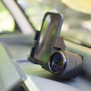 Op de afbeelding: Een zwarte telefoonhouder met een blauwe meter eraan bevestigd. De telefoonhouder is gemonteerd op het dashboard van een auto.