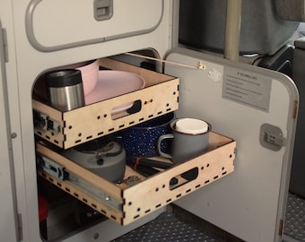Könnte beinhalten: Zwei Holzkisten mit Auszugsschienen sind geöffnet und zeigen eine Auswahl an Küchenutensilien, darunter eine rosa Schüssel, eine graue Tasse, ein blauer Emailletopf und eine silberne Thermoskanne. Die Schubladen sind aus hellem Holz mit einer dunkelbraunen Beize und haben eine Reihe kleiner Löcher an den Rändern.