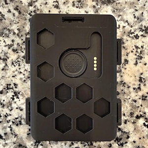 May include: A black rectangular device with a series of hexagonal cutouts and a circular button. The device has a small, raised section with four yellow dots and two side clips. The object is likely a tech accessory.