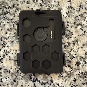 Könnte beinhalten: Schwarzes rechteckiges Gerät mit einer Reihe sechseckiger Aussparungen und einem kreisförmigen Knopf. Das Gerät hat einen kleinen Clip oben und vier goldfarbene Punkte an der Seite. Das Gerät besteht aus einem mattschwarzen Material.