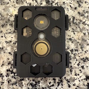 Peut inclure: Un appareil rectangulaire noir avec un dessus articulé et de multiples découpes hexagonales. L'appareil comporte un composant circulaire de couleur or et un composant circulaire plus petit et transparent. L'appareil repose sur une surface mouchetée.