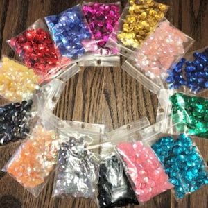 May include: Assortment of 14 small bags of colourful sequins in various colours, including red, blue, green, pink, gold, and black. The sequins are packaged in clear plastic bags.