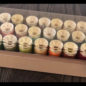 May include: A clear plastic box with a beige lid filled with 24 spools of thread. The spools are various colors, including red, green, blue, and yellow. The spools are labeled with the text "Gutermann 100 m."