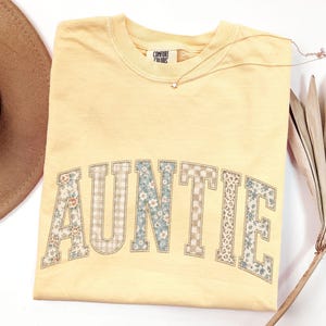 May include: A butter yellow short-sleeve t-shirt with a crew neckline and a Comfort Colors brand label. The front features the word AUNTIE in large block letters, each filled with a different pattern including floral, gingham, and checkered prints.