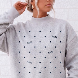 May include: A light grey crewneck sweatshirt featuring a repeating pattern of small dark navy blue polka dots interspersed with the word "MAMA" written in a dark navy blue script font across the front of the garment.