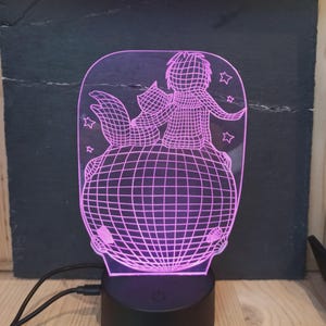 Può includere: Lampada da notte decorativa con un design a filo rosa raffigurante un personaggio e una volpe su un globo. La lampada, con base nera e cavo USB, è un elemento decorativo.