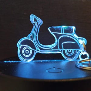 Op de afbeelding: Een heldere acryl scooter sleutelhanger met een blauwe neon omtrek. De sleutelhanger heeft een gedetailleerd ontwerp van een vintage scooter, compleet met wielen, zitting en stuur. De sleutelhanger is bevestigd aan een zilveren metalen ring.