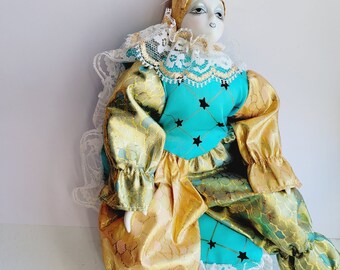 May include: A porcelain doll dressed as a jester with a blue and gold costume. The doll has a white face with painted black eyes and pink lips. The doll is wearing a white lace collar and a pink flower on its head.