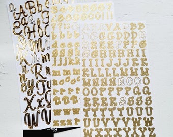 May include: Three sheets of gold alphabet and number stickers. The first sheet has lowercase letters in a cursive font. The second sheet has numbers and punctuation marks. The third sheet has uppercase letters in a block font.