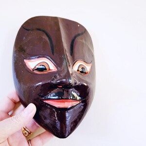 May include: A dark brown wooden mask with painted details. The mask has a wide, smiling mouth with pink lips, black eyes with white and orange accents, and black eyebrows.