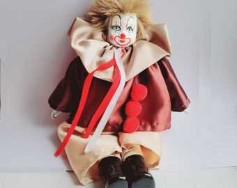 May include: A vintage porcelain clown doll with a painted face, wearing a satin costume in shades of brown, beige, and red. The doll has a fluffy, light brown wig, a large collar, and red and white ribbons. The clown's shoes are black.