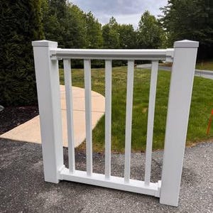 May include: A white vinyl railing section featuring two square vertical posts connected by a horizontal top rail and a bottom rail with five evenly spaced vertical balusters between them, creating a clean and structured architectural barrier.