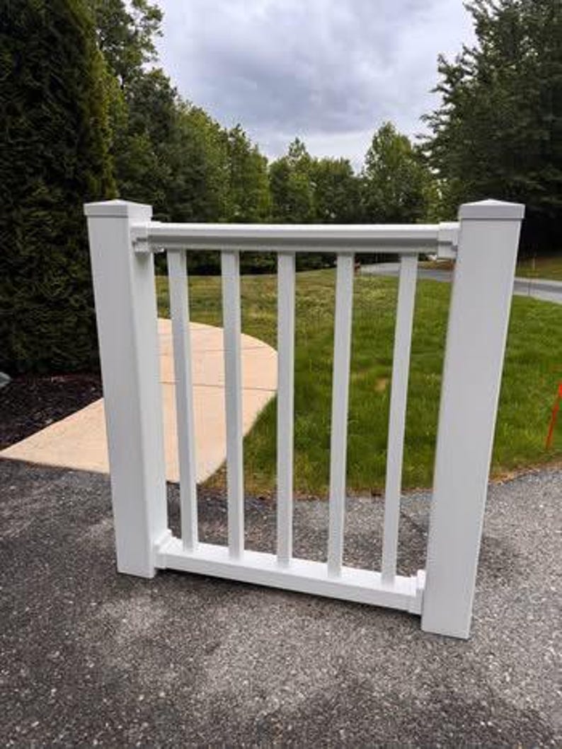 May include: A white vinyl railing section featuring two square vertical posts connected by a horizontal top rail and a bottom rail with five evenly spaced vertical balusters between them, creating a clean and structured architectural barrier.