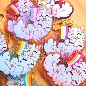 Peut inclure: Plusieurs porte-clés scintillants en forme de chat. Chaque porte-clé représente un chat blanc tenant un drapeau aux couleurs arc-en-ciel ou aux couleurs du drapeau de la fierté. Les porte-clés ont une attache dorée et sont entourés d'une bordure transparente en forme d'étoile.