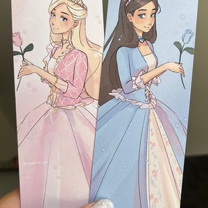 Op de afbeelding: Twee geïllustreerde bladwijzers met prinsessen in baljurken. De prinses links is blond met een roze jurk en een roos. De prinses rechts heeft lang bruin haar en een blauwe jurk met een blauwe roos.