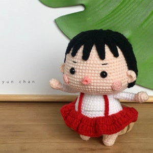 May include: A handmade crochet doll of a girl with black hair, wearing a white shirt with red suspenders and a red skirt. The doll has a neutral expression and is standing on a wooden surface. The text "Jaeyun chan" is visible.