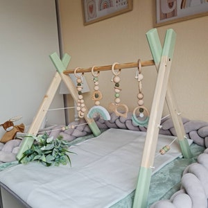 Könnte beinhalten: Hölzerner Baby-Spielbogen mit grün-weißer Lackierung. Der Spielbogen hat eine Holzstange mit fünf hängenden Spielzeugen, darunter ein Regenbogen, ein Holzring, eine Holzperlenkette und zwei Holzbeißringe. Der Spielbogen ist auf einer weiß-grünen Spielmatte aufgebaut.