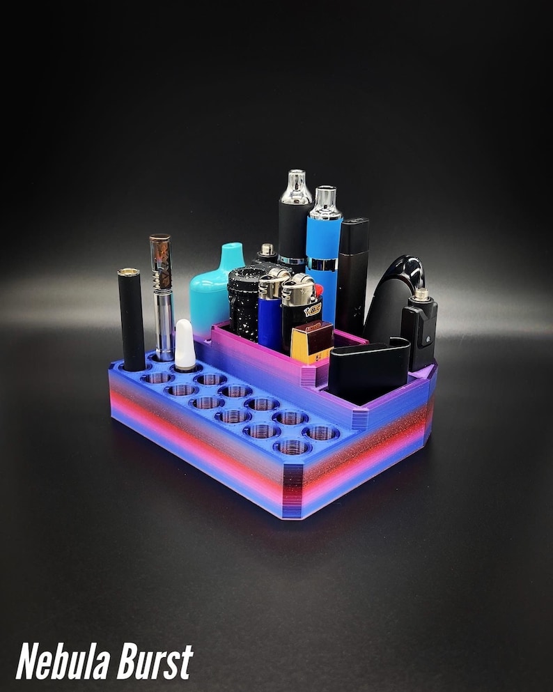 May include: A multi-colored, 3D-printed organizer with a gradient of blue, red, and purple. It holds various vaping devices, lighters, and accessories. The text "Nebula Burst" is printed at the bottom.