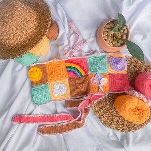 May include: A colorful crocheted top with a rainbow, smiley face, cloud, and bear design. The top is made of different colored squares and has a pink and brown tie.