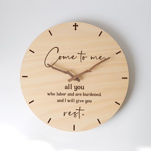 May include: A circular wooden clock with a light wood grain. The clock face has the text "Come to me, all you who labor and are burdened, and I will give you rest." The clock hands are made of small twigs.