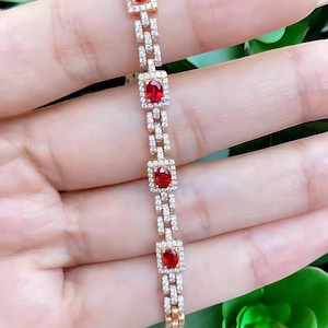 Puede incluir: Una pulsera de oro rosa con piedras rojas rectangulares engastadas en marcos cuadrados, conectadas por eslabones más pequeños con piedras claras de acento. La pulsera se muestra sobre un fondo suave, resaltando su elegante diseño.