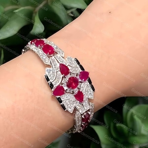 May include: A silver-toned bracelet featuring a central floral-inspired cluster of pear-shaped and round deep red gemstones, surrounded by small clear pavé-set stones and accented with black enamel details along the articulated metal links.