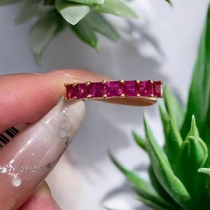 May include: A gold ring featuring a series of vibrant, square-cut, ruby-colored gemstones. The ring's design showcases a continuous line of the gemstones, set closely together. The ring is held against a backdrop of green plants and a white surface.