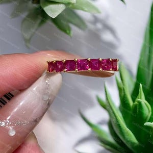 May include: A gold band ring featuring a row of seven square-cut, vibrant red rubies set in a shared-prong setting. The stones are aligned horizontally, showcasing a deep crimson hue and faceted surfaces that reflect light across the metal band.