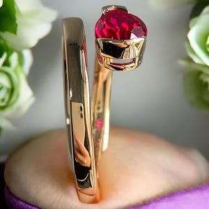 Puede incluir: Un anillo dorado con una gran gema roja facetada. El anillo tiene un diseño abierto único que se envuelve alrededor del dedo. La gema está engastada en un bisel dorado, sobre un fondo morado suave.