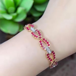 Puede incluir: Un brazalete dorado con un diseño de eslabones de cadena. Cada eslabón está adornado con pequeñas gemas rojas vibrantes. Se intercalan eslabones con gemas blancas. El brazalete se muestra en una muñeca.