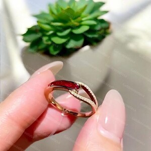 May include: A gold-toned ring featuring a deep red, rectangular-cut stone set horizontally on one side of the band. The band has a split design, with one side encrusted with small, clear, sparkling stones that meet the red stone at the center.