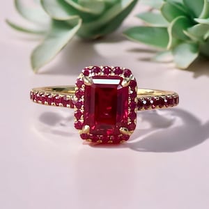 May include: A gold ring with a large, rectangular, ruby-colored center stone, surrounded by smaller red gemstones. The ring's band is also adorned with small red stones. The ring is set against a soft pink background, with green foliage in the background.
