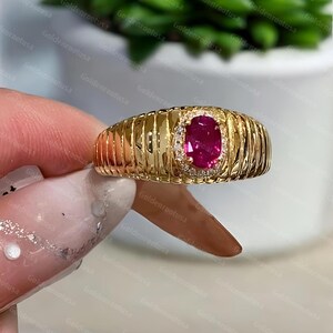 Könnte beinhalten: Ein Goldring mit einem zentralen, oval geschliffenen, leuchtend pink-roten Edelstein, der von einem Kranz aus kleinen, klaren Steinen umgeben ist. Die Ringschiene hat eine breite, polierte Goldoberfläche mit vertikalen Rillenstrukturen.
