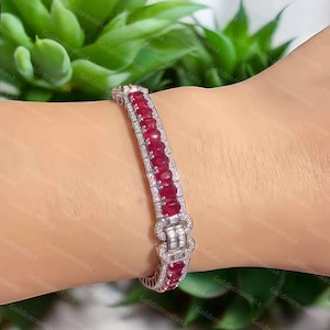 May include: A silver-toned bracelet featuring a central row of oval-cut red rubies flanked by two rows of small clear stones. The design includes a rectangular clasp section adorned with baguette-cut clear stones and a decorative border of small clear stones.