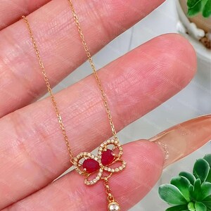 May include: A gold chain necklace featuring a butterfly-shaped pendant. The pendant is adorned with two pear-cut red gemstones and small clear stones along the wings. A single clear stone hangs from the bottom of the butterfly design.