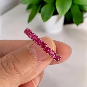 Puede incluir: Un anillo de oro con una banda de gemas rosas vibrantes. Las gemas están muy juntas, creando una línea continua de color. El anillo se sostiene en una mano, con un fondo borroso de follaje verde.