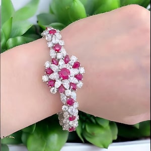 May include: A silver bracelet with a floral design, featuring ruby-red and clear gemstones. The bracelet's central flower-shaped cluster is surrounded by smaller gemstones, creating a sparkling, elegant accessory. The bracelet is worn on a wrist.