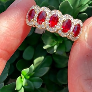 May include: A gold ring with five oval, deep red gemstones, each encircled by a halo of small, brilliant white diamonds. The ring is held up against a background of green leaves.