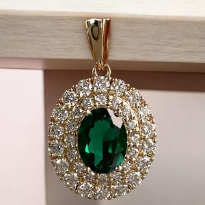 Pode incluir: Pingente de ouro com uma grande esmeralda oval, rodeada por um duplo halo de pedras brancas brilhantes. O pingente tem uma argola de ouro para prender a uma corrente.