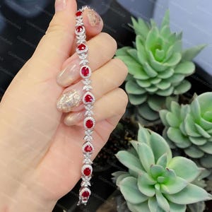 Peut inclure: Un bracelet argenté composé d'une alternance de pierres précieuses rouges ovales et en forme de cœur. Chaque pierre est entourée d'un halo de petites pierres claires, avec des maillons en forme de feuille sertis de pierres claires.
