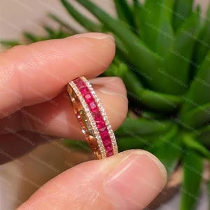 Op de afbeelding: Een roségouden ring met een centrale rij vierkant geslepen, levendig rode robijnen, geflankeerd door twee delicate rijen kleine, heldere pavé-gezette diamanten. De ring heeft een gepolijste metalen afwerking en een kanaalzetting voor de stenen.